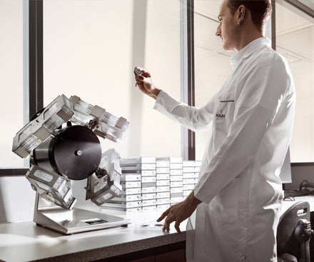 A man in a lab coat inspects a watch near a device, with stacked boxes on the counter by a window.