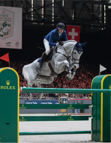 A rider in blue jumps a gray horse over a green Rolex obstacle at an indoor equestrian event.