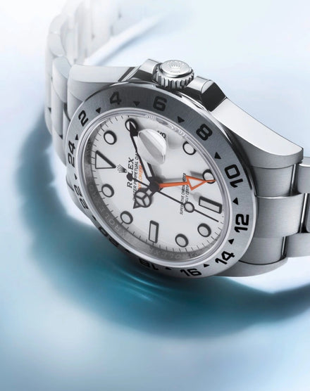A silver Rolex watch with a white face and orange hand, displayed on a light blue surface.