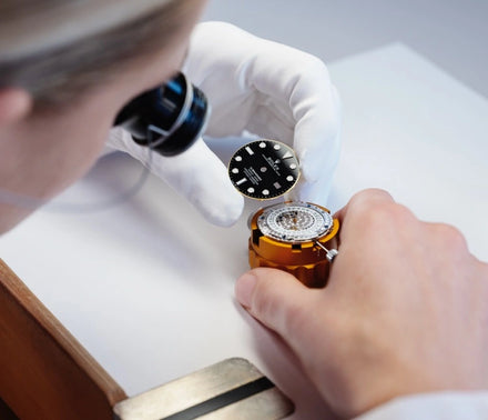 A watchmaker in white gloves examines a black watch face and works on a watch mechanism.