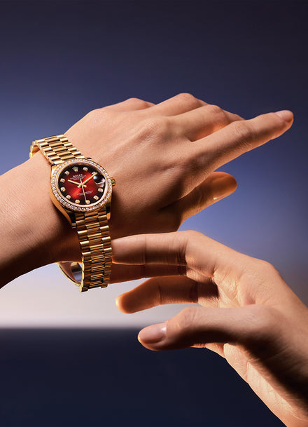 A gold wristwatch with a red face and diamond bezel on a persons wrist against a blurred background.