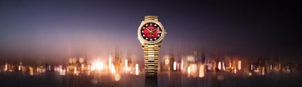 A gold wristwatch with a red face and diamond markers, set against a blurred city lights background.