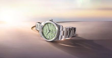 A silver wristwatch with a light green dial rests on a smooth, curved surface in soft lighting.