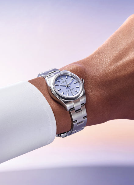 A silver wristwatch with a white face is worn on a wrist with a white shirt cuff against a soft gradient background.