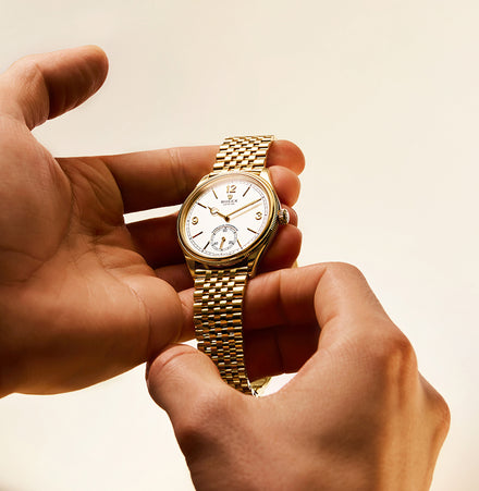 A person’s hands holding a gold wristwatch with a white dial against a beige background.
