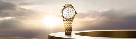 Gold Rolex watch with a white dial displayed against a bright, cloudy sky and golden circular surface.