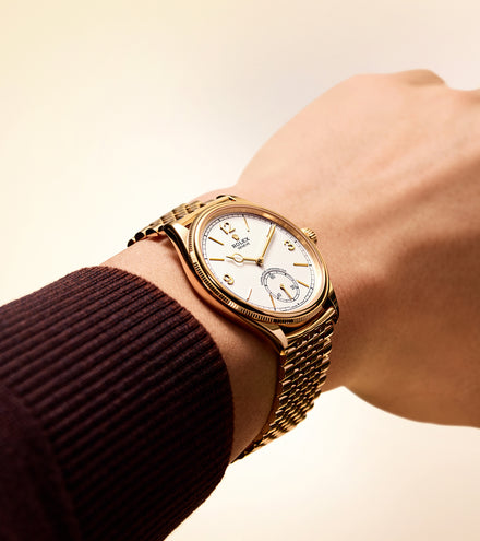 A gold wristwatch with a white dial is worn on a persons wrist in a brown sweater.