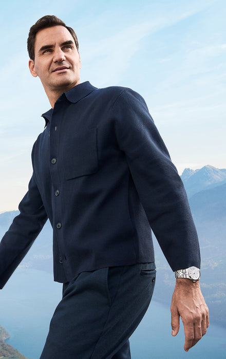 A man in a navy jacket stands outdoors with mountains and a lake in the background.