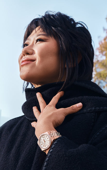 Woman in a black coat looking up, hand on chest, wearing a gold wristwatch, outdoors with blue sky.