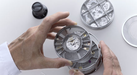 Hands holding a round mesh tray with small watch parts, with similar trays and a magnifier on a white surface.