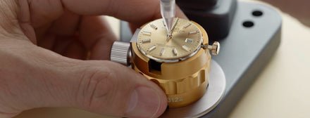 A close-up of hands assembling a gold wristwatch with a precision tool.