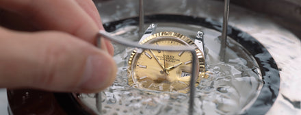 A hand dips a gold-faced Rolex watch into water, partially submerging it during a cleaning process.