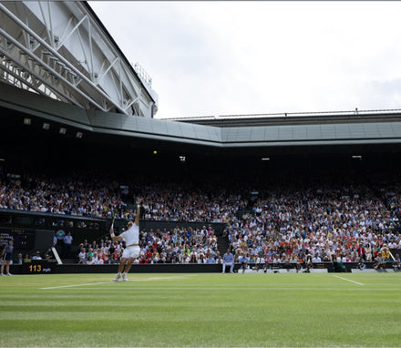 A tennis player serves on a grass court at a stadium filled with spectators under a partly cloudy sky.