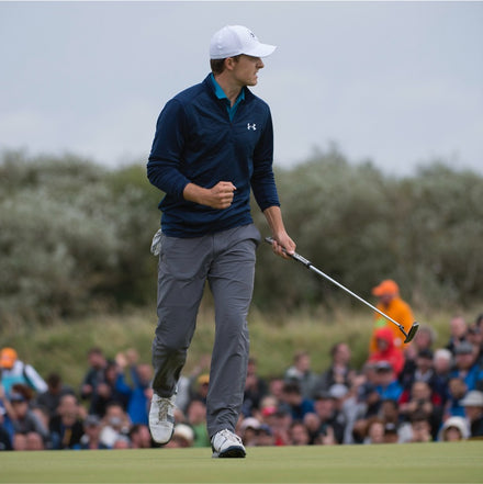 Golfer in a navy jacket celebrates on the green, clenching his fist, with a crowd watching in the background.