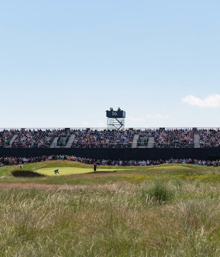 A golfer prepares to putt on a green, watched by a large crowd in stadium seating at hole 16.