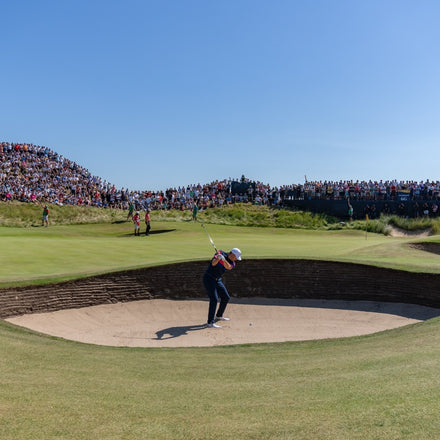 Golfer hitting a shot from a sand bunker on a sunny day, with a large crowd watching in the background.