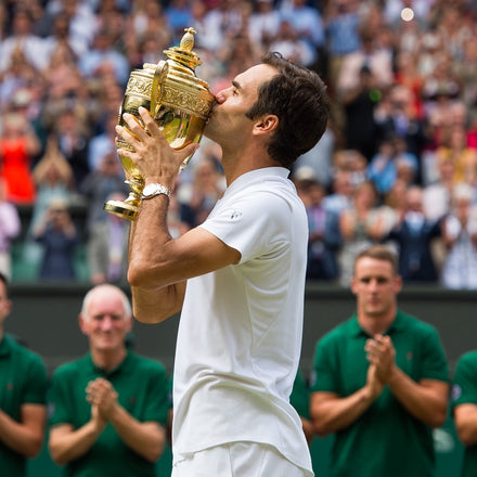 A tennis player in white kisses a gold trophy as people applaud in the background on a tennis court.