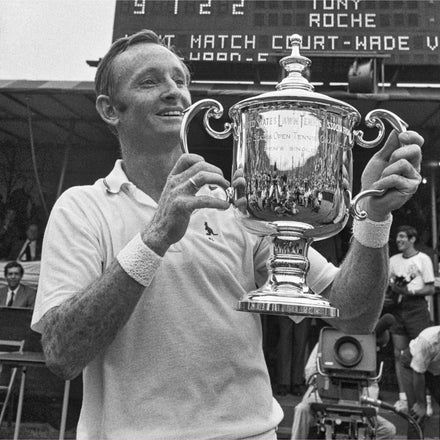 A smiling tennis player holds up a large trophy on a court with a scoreboard in the background.