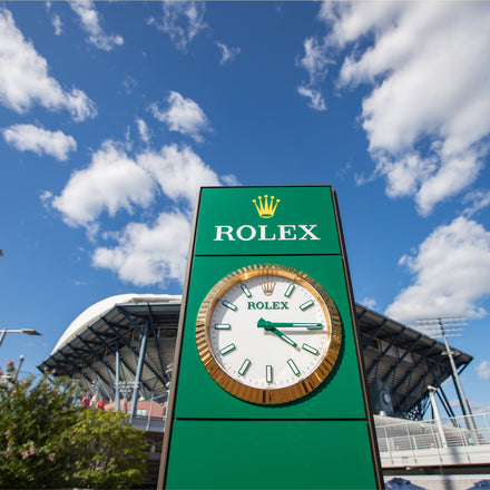 Green Rolex clock stands outdoors with a stadium and blue sky with clouds in the background.