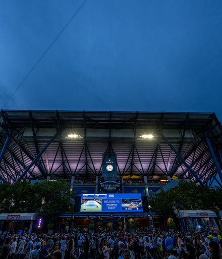 Crowd gathers outside Arthur Ashe Stadium at dusk with large screen displays and a clock above the entrance.
