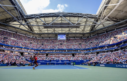 Wide view of a crowded tennis stadium during a match, with players on the court and a partly open roof above.