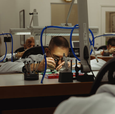 A man in a lab coat inspects a small object at a workbench with tools and magnifying equipment.