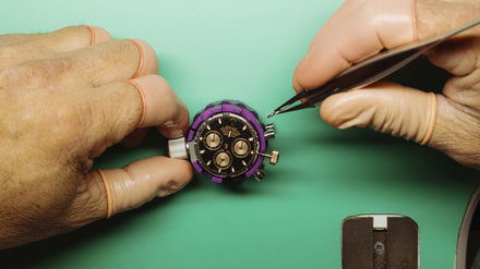 Gloved hands working on a wristwatch with tools on a green surface.