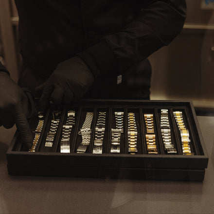 A person wearing black gloves displays a tray of various metal watch bands on a counter.
