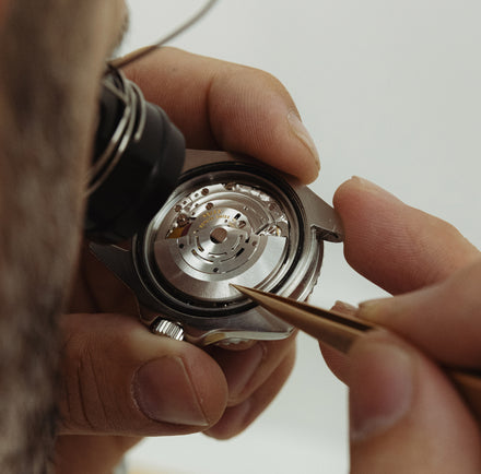 A watchmaker uses tweezers to adjust the intricate mechanism inside a wristwatch case.