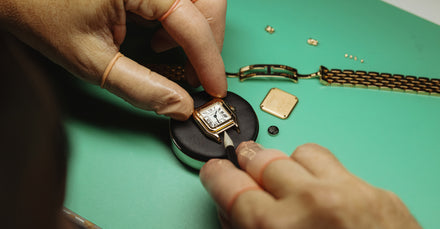 A watchmaker repairs a gold wristwatch on a green surface with tools and watch parts nearby.