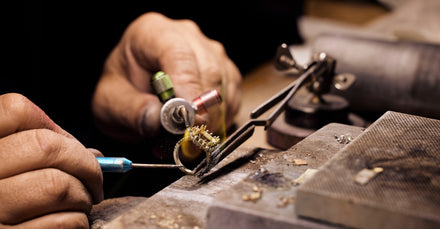 Hands crafting jewelry with tools, holding a ring and working on its intricate details at a workbench.