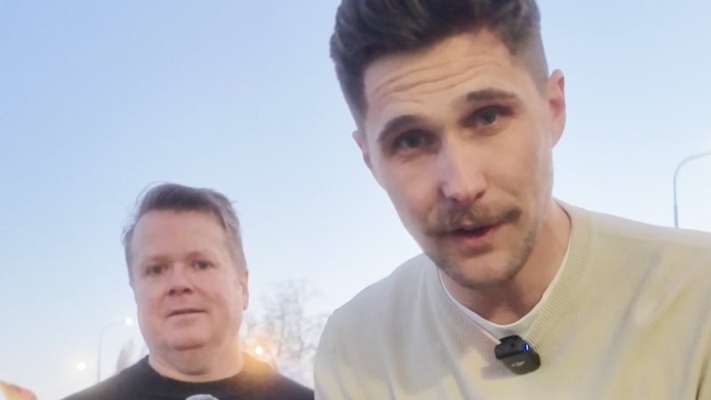 Two men standing outdoors at dusk, one looking directly at the camera, both wearing microphones.