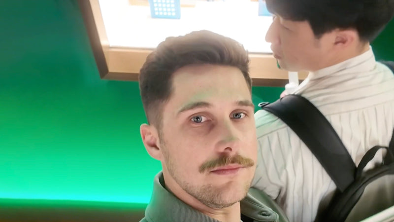 Two men stand indoors; one faces the camera, and the other faces away, both near a green wall and display.