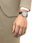 Round watch with white dial, gold accents, and dark leather strap worn on wrist with beige suit jacket.