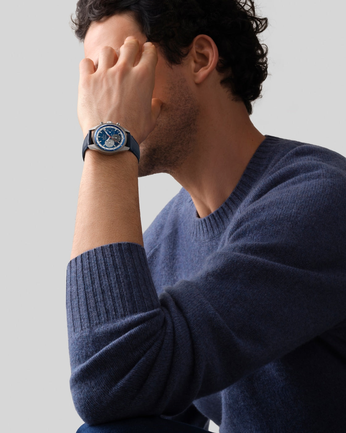 A man in a blue sweater covers part of his face with his hand, showcasing the Zenith Chronomaster Original blue-faced wristwatch.