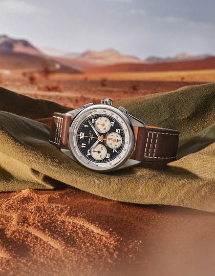 A wristwatch with a silver-toned case, black dial, and brown leather strap rests on draped olive-green fabric in a desert landscape with reddish sand and distant mountains.
