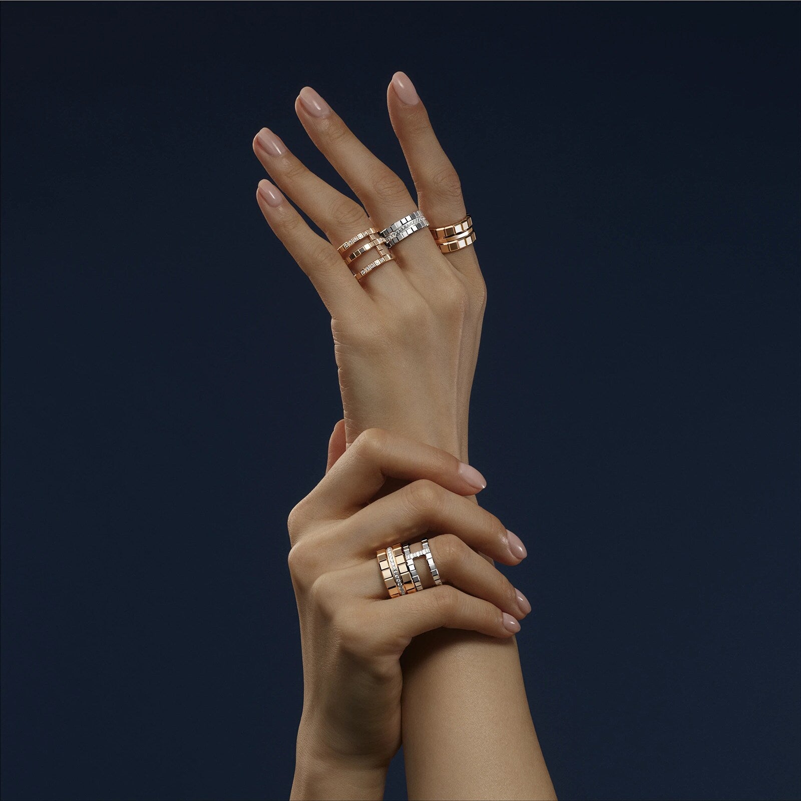 Two hands with manicured nails, each adorned with Chopards Ice Cube diamond ring and other gold and silver rings, are crossed against a dark background, showcasing minimalist elegance and a modern aesthetic.