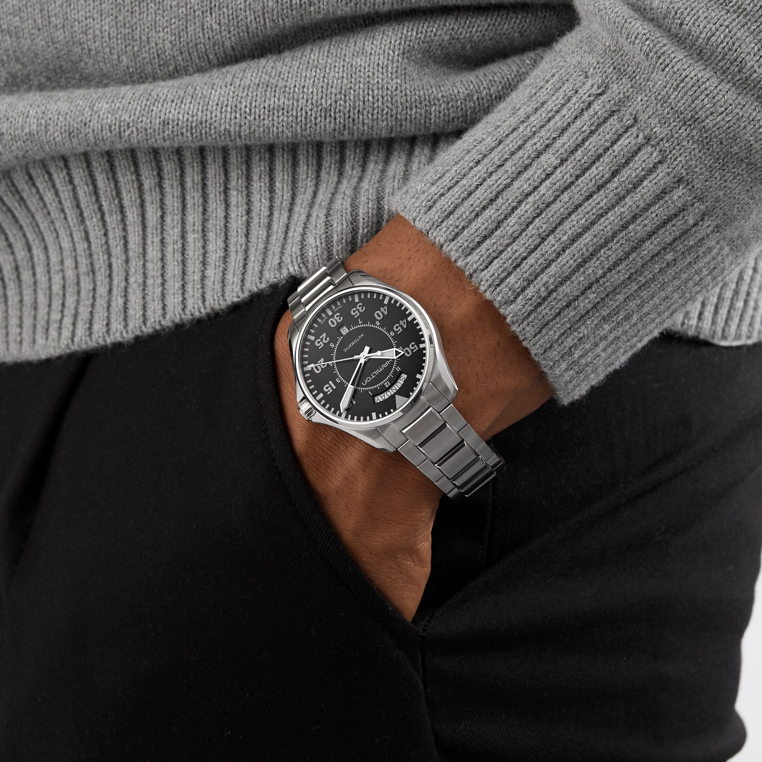 A person in a gray sweater and black pants has their hand in their pocket, showing off the Hamilton Khaki Aviation Pilot Day Date Auto, a vintage-inspired watch with automatic movement and a sleek black dial.