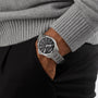 A person in a gray sweater and black pants has their hand in their pocket, showing off the Hamilton Khaki Aviation Pilot Day Date Auto, a vintage-inspired watch with automatic movement and a sleek black dial.
