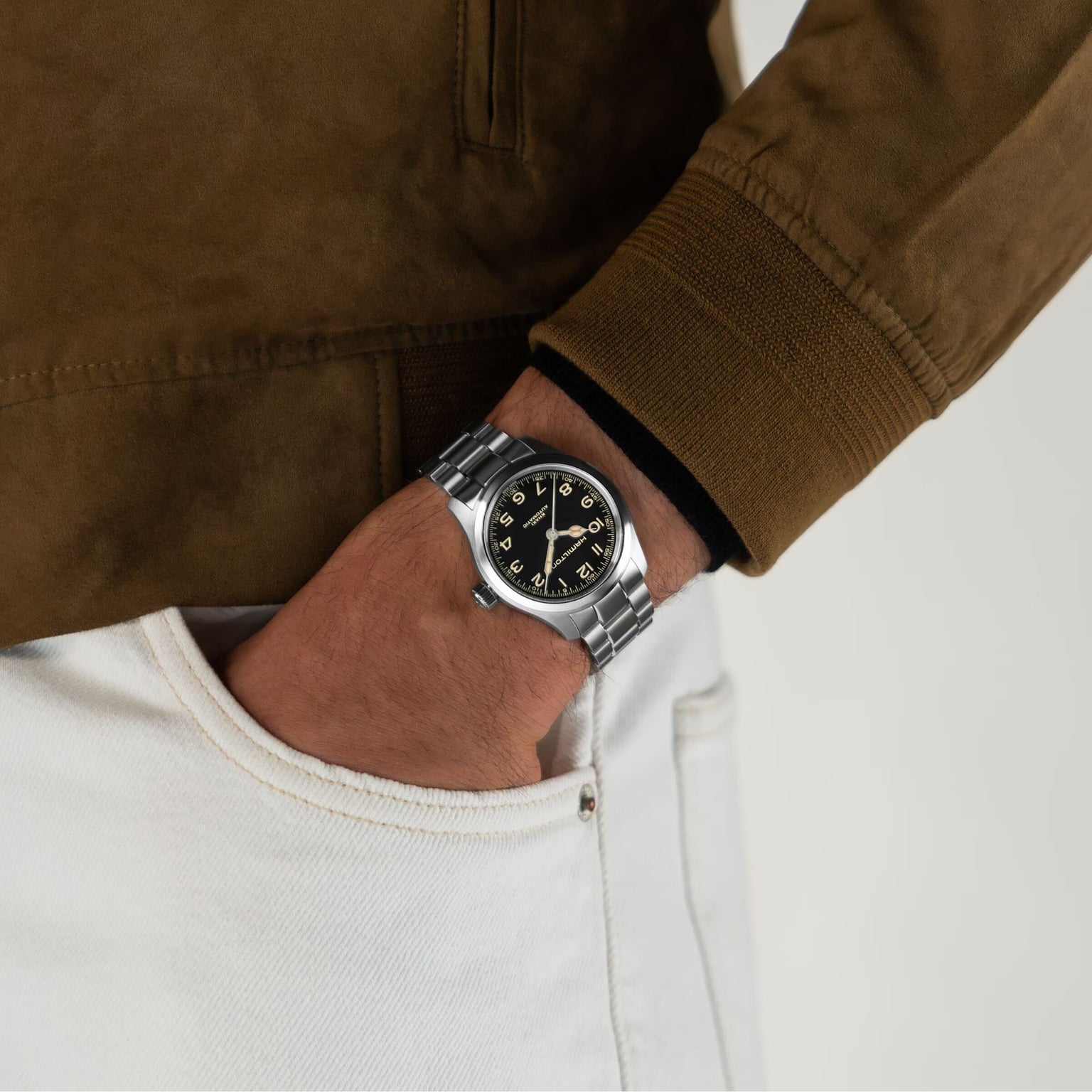 A person in a brown suede jacket and white pants keeps their left hand in their pocket, revealing a vintage-style Hamilton Khaki Field Murph 38mm watch with a black dial and Arabic numerals.