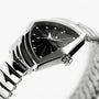 A close-up of the Hamilton Ventura S Quartz shows its silver case, black triangular dial with metallic hour markers, zigzag pattern, and a sleek metal link band inspired by Mid-Century Modern design.