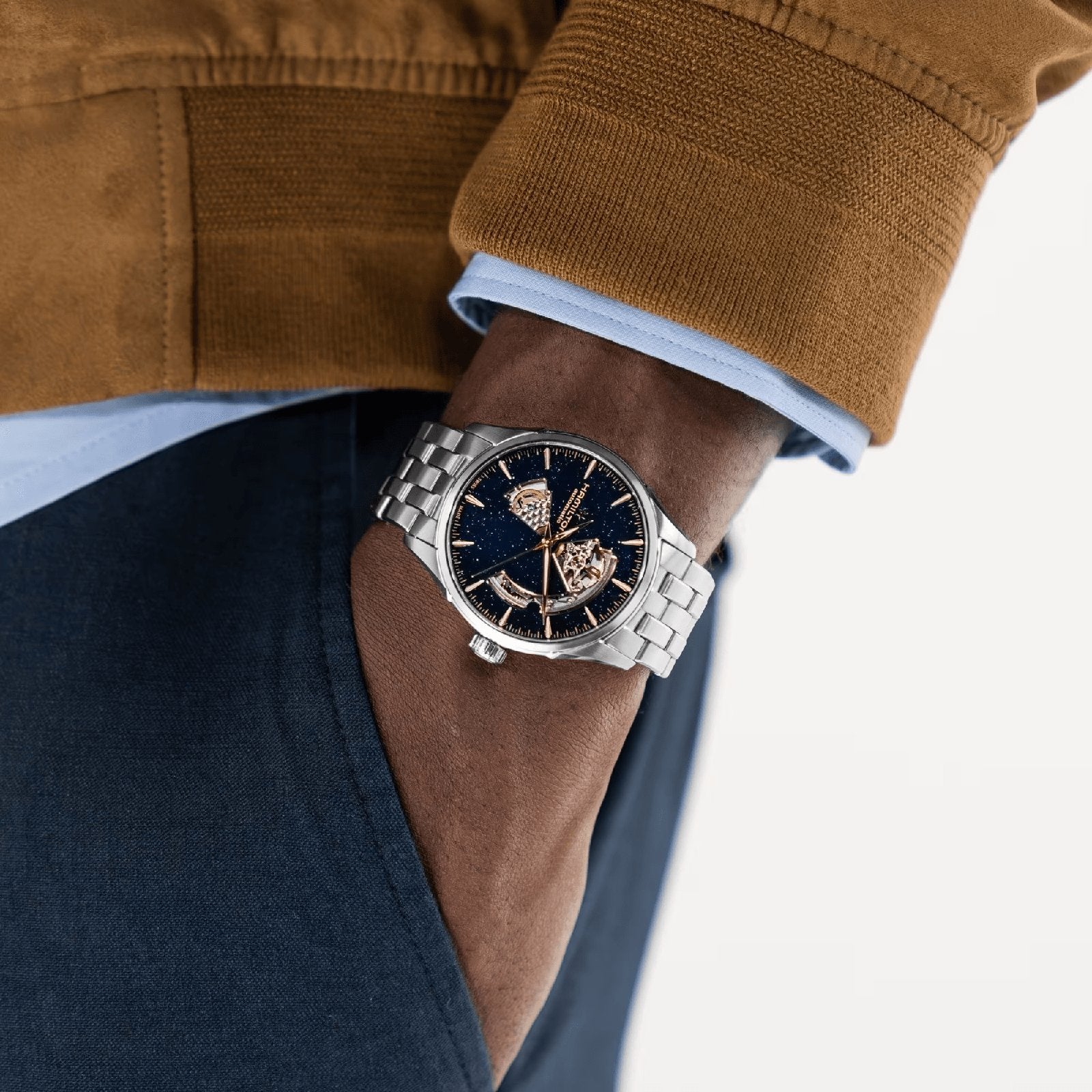 A person in a brown jacket and blue pants wears the Hamilton Jazzmaster Open Heart Auto 42mm, displaying its dark openwork dial and automatic movement, with their hand in their pocket.