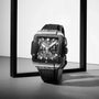 The Hublot Square Bang Unico Titanium Ceramic watch with a black rubber strap stands upright in a minimalist black frame on a gray surface, showing its intricate UNICO movement through the sapphire crystal.