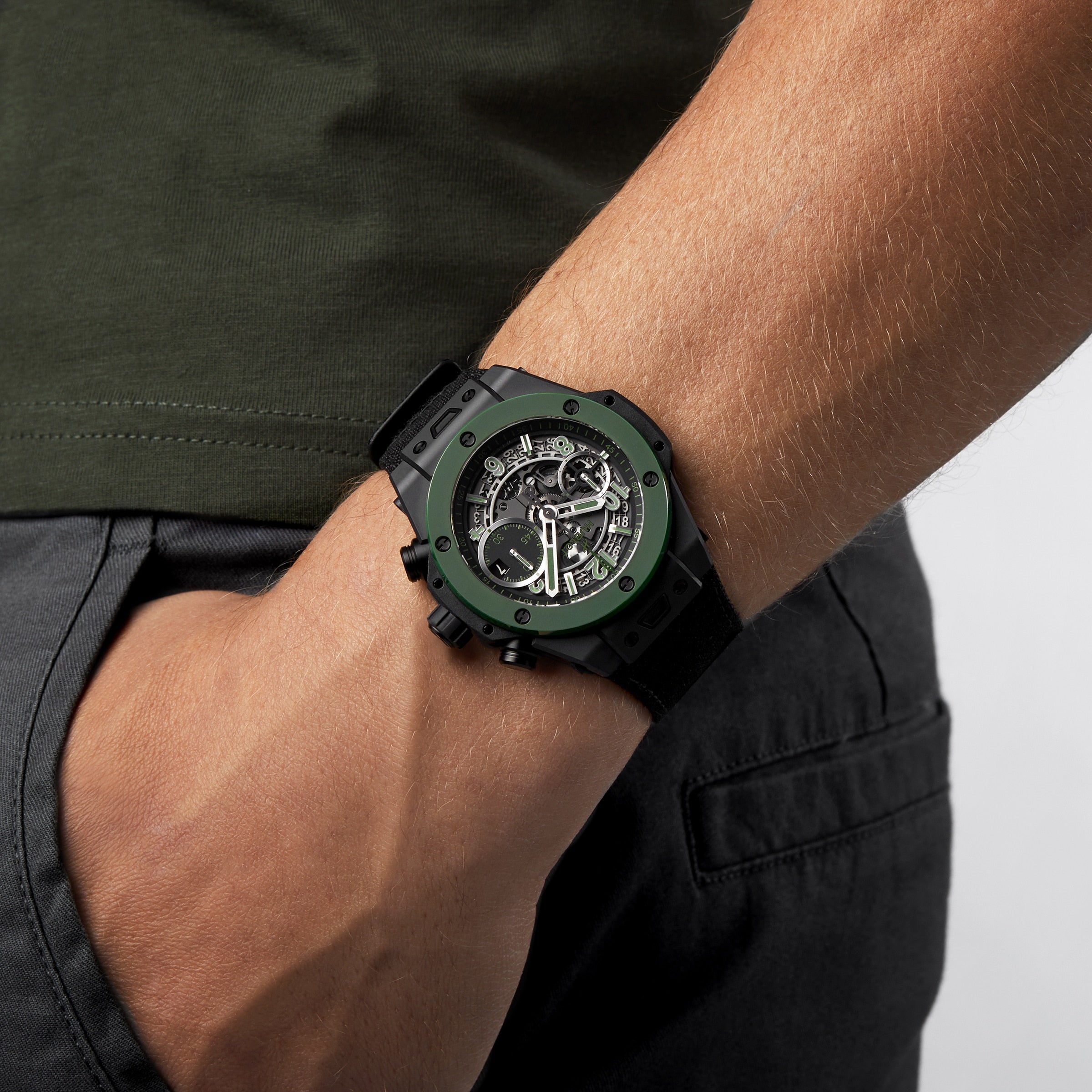 A person in a dark green shirt and black pants wears the Hublot Big Bang Unico The Watches of Switzerland Group Exclusive with a green bezel on their left wrist. Their hand rests casually in their pocket.