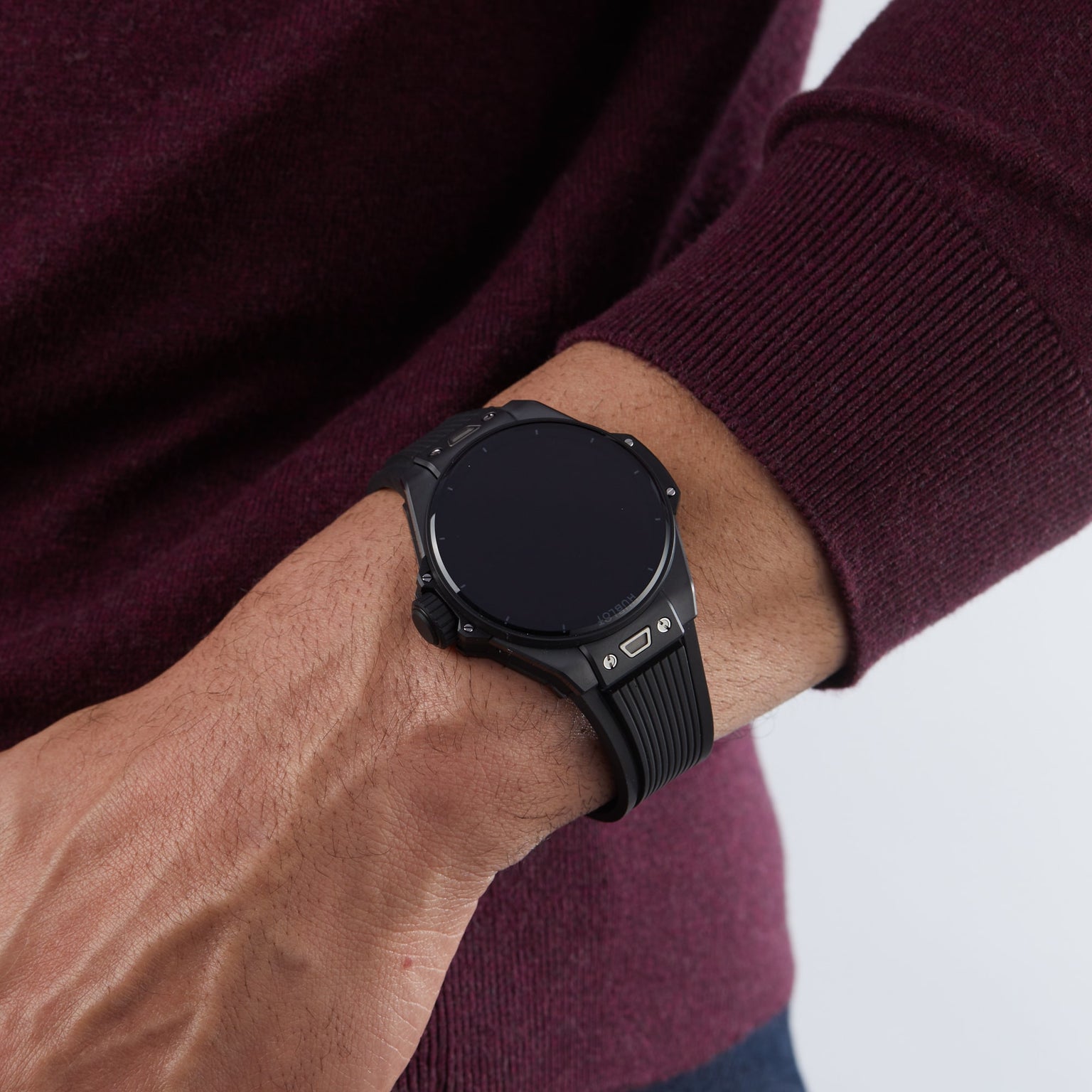 A person wearing a Hublot Big Bang E Gen3 Black Magic smartwatch with a blank screen on their left wrist, paired with a maroon long-sleeve sweater.