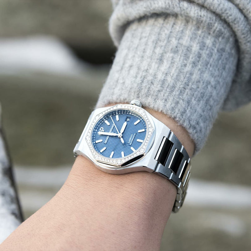 A Girard-Perregaux Laureato 34 silver wristwatch with a blue dial and diamond-set octagonal bezel is shown on a wrist with a gray knit sleeve.