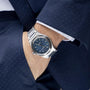 A person in a navy suit wears the Girard-Perregaux Laureato 42, a silver wristwatch with a blue dial, with their hand in their pocket.