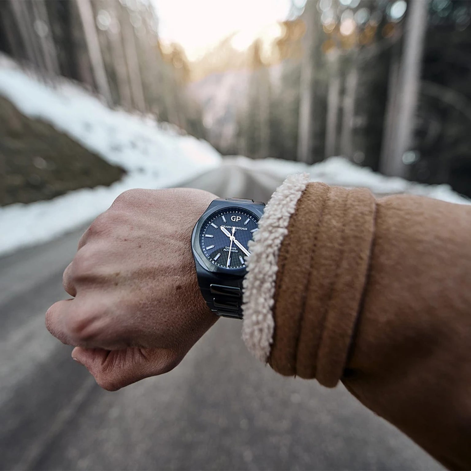A person in a brown coat displays their Girard-Perregaux Laureato 42, featuring an octagonal bezel, while standing on a road lined with snowy trees against a blurred forest backdrop.