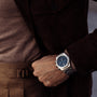 A person in a brown outfit showcases a close-up of their hand and waist, wearing a Girard-Perregaux Laureato Chronograph 42 with an octagonal bezel and blue dial.