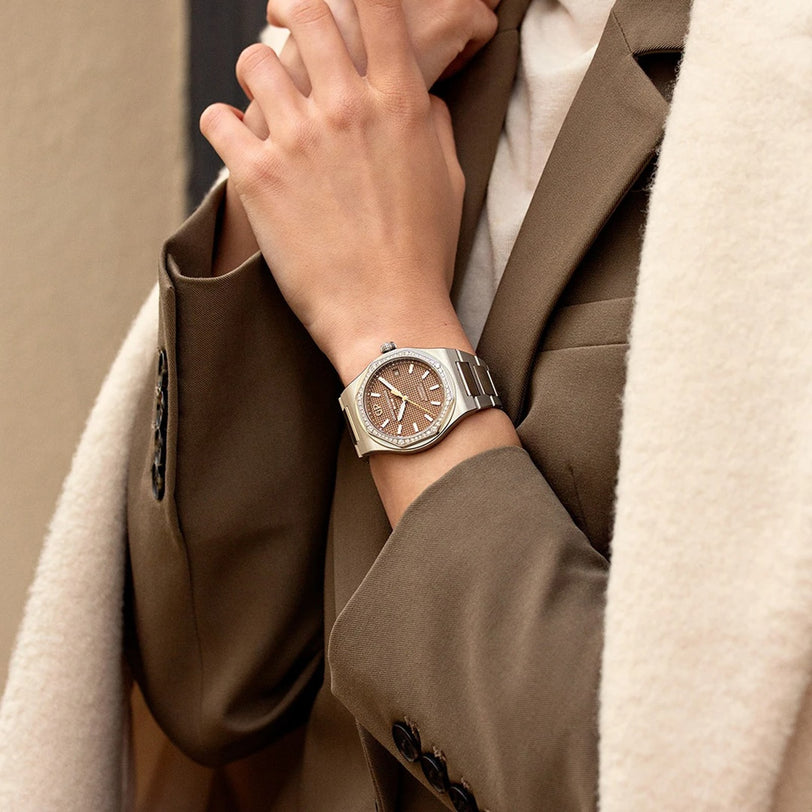 Person wearing a brown blazer and beige coat, displaying a Girard-Perregaux Laureato 38 with a silver case, brown dial, and octagonal bezel on their left wrist.